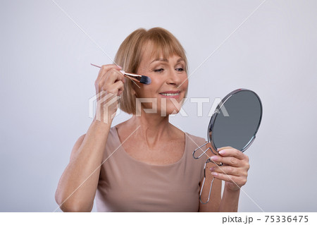 Lovely senior lady using cosmetic brush to apply decorative makeup on her face, looking in mirror on light background Lovely senior lady using cosmetic brush to apply decorative makeup on her face, looking in mirror on light background 75336475
