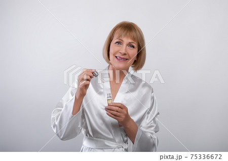 Cheerful senior lady in dressing gown using face serum on light studio background. Skincare and wellness Cheerful senior lady in dressing gown using face serum on light studio background. Skincare and wellness 75336672