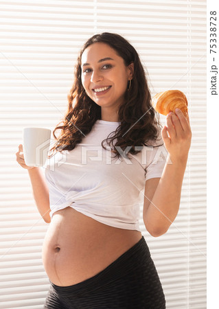 Beautiful pregnant woman holding croissant and cup of coffee in her hands during morning breakfast. Concept of good health and positive attitude while expecting baby 75338728