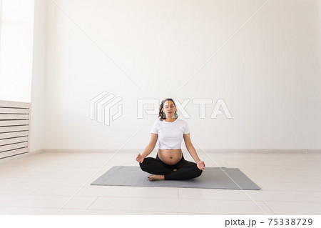Healthy beautiful pregnant woman meditates before yoga class and relaxes while sitting on mat on the floor. Concept of physical and mental preparation of body for childbirth. Copyspace 75338729