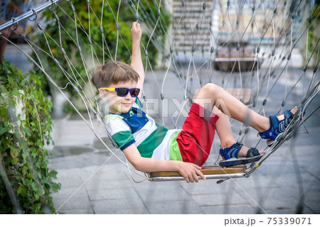 Little adorable kid sitting on chain swing during summer holidays on vacation. Boy relaxed and happy wearing casual clothes in sunglasses. Active summer leisure for kids in the city concept Little adorable kid sitting on chain swing during summer holidays on vacation. Boy relaxed and happy wearing casual clothes in sunglasses. Active summer leisure for kids in the city concept 75339071