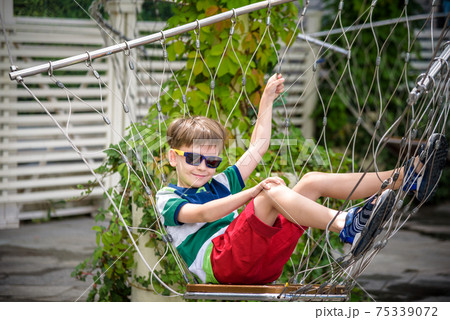 Little adorable kid sitting on chain swing during summer holidays on vacation. Boy relaxed and happy wearing casual clothes in sunglasses. Active summer leisure for kids in the city concept Little adorable kid sitting on chain swing during summer holidays on vacation. Boy relaxed and happy wearing casual clothes in sunglasses. Active summer leisure for kids in the city concept 75339072