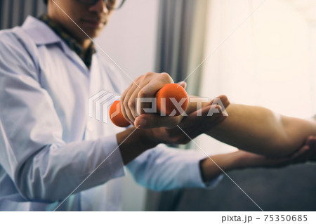Asian physiotherapist helping a patient lifting dumbbells work through his recovery with weights in clinic room. 75350685