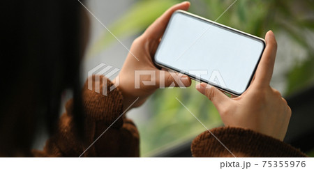 Cropped image of beautiful woman on brown cotton sweater holding a white blank screen smartphone in her hands while sitting and relaxing on her holidays in comfortable sitting room as background. Cropped image of beautiful woman on brown cotton sweater holding a white blank screen smartphone in her hands while sitting and relaxing on her holidays in comfortable sitting room as background. 75355976