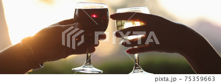 Male and female hands are holding glass of red and white wine against sunset background. 75359663