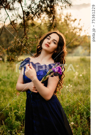 Party graduation prom fairytale concept. Beautiful brunette young woman in blue prom dress with iris flower on nature background. Outdoor romantic portrait of brunette girl in long evening dress Party graduation prom fairytale concept. Beautiful brunette young woman in blue prom dress with iris flower on nature background. Outdoor romantic portrait of brunette girl in long evening dress 75361229
