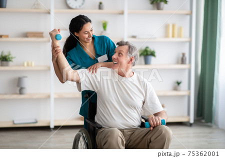 Rehabilitation of disabled people. Young physiotherapist helping senior male patient in wheelchair exercise at home 75362001