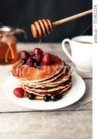 Pancakes with berries and honey on a white plate, wooden spoon, jar, coffee cup Pancakes with berries and honey on a white plate, wooden spoon, jar, coffee cup 75362226