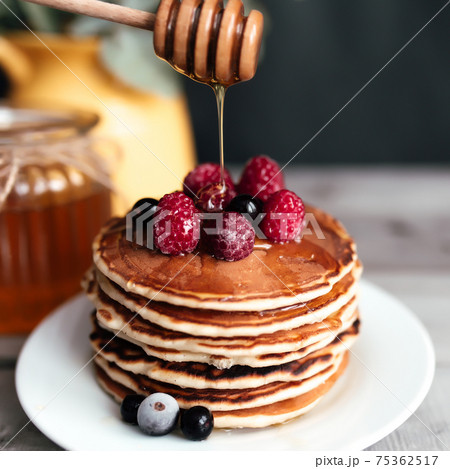 Juicy pancakes with berries and honey on a white plate, spoon, jar, wooden table 75362517
