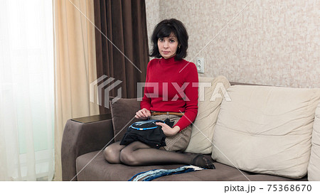 Woman in red clothes sitting on the couch is embroidering a picture in a cross Woman in red clothes sitting on the couch is embroidering a picture in a cross 75368670