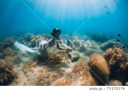 Freediver woman glides with sand in hand. Free diver with fins posing underwater in blue ocean Freediver woman glides with sand in hand. Free diver with fins posing underwater in blue ocean 75370174