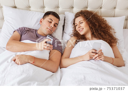 Happy affectionate couple spend morning time in bedroom, being always in touch, use modern gadgets for surfing internet and online communication. Family with mobile phones in comfortable bed 75371037