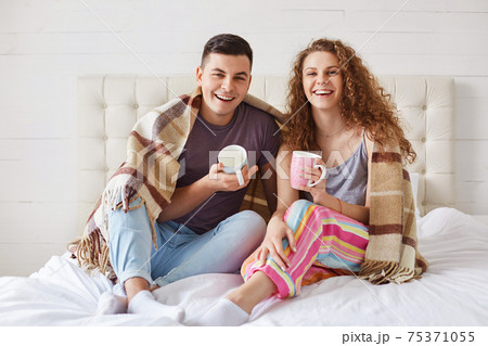 Positive young family couple wear pyjamas, drink coffee in morning at bedroom, enjoy starting new day. Affectionate female and make enjoy togetherness at cozy domestic atmosphere. Family concept Positive young family couple wear pyjamas, drink coffee in morning at bedroom, enjoy starting new day. Affectionate female and make enjoy togetherness at cozy domestic atmosphere. Family concept 75371055