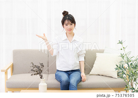 部屋のソファーに座って案内のジェスチャーをする若い女性 75376505