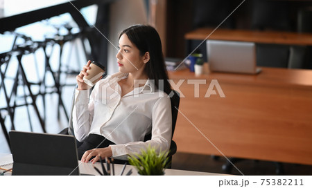 Attractive businesswoman relaxing at her workplace and looking away during a coffee break. Attractive businesswoman relaxing at her workplace and looking away during a coffee break. 75382211
