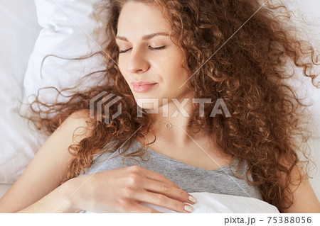 Close up shot of pleasant looking female naps in comfortable bed alone, keeps eyes closed, has curly hair, wears casual domestic clothing, enjoys good rest. People, rest and lifestyle concept Close up shot of pleasant looking female naps in comfortable bed alone, keeps eyes closed, has curly hair, wears casual domestic clothing, enjoys good rest. People, rest and lifestyle concept 75388056