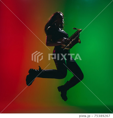 Silhouette of young girl with saxophone posing isolated on green-red background. Silhouette of young girl with saxophone posing isolated on green-red background. 75389267