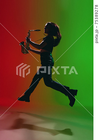 Silhouette of young girl with saxophone posing isolated on green-red background. Silhouette of young girl with saxophone posing isolated on green-red background. 75389278