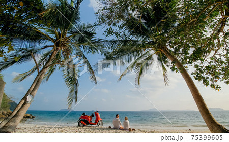 Scooter road trip. Lovely couple on red motorbike in white clothes on sand beach. People walking near the tropical palm trees, sea. Motorcycle rent. 75399605