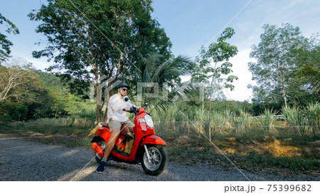 Man on red motorbike in white clothes drive on forest road trail trip. One men caucasian tourist go on scooter, nearby tropical palm tree. 75399682