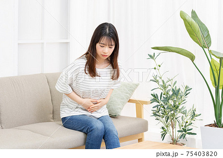 部屋のソファーに座ってお腹に手を当てる若い女性 75403820