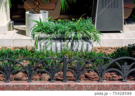 stone flower pot with flowers in a landscape bed with bushes and trees sprinkled with tree bark mulching landscape of the backyard of a summer cottage with black iron fence, nobody. stone flower pot with flowers in a landscape bed with bushes and trees sprinkled with tree bark mulching landscape of the backyard of a summer cottage with black iron fence, nobody. 75428599