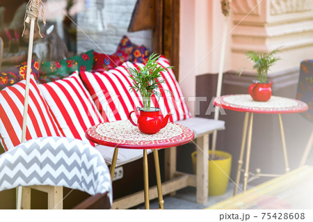 a red round table with a tablecloth and a flowerpot in the form of a teapot with a green plant by a chair with pillows, an outdoor cafe with empty seats, nobody. a red round table with a tablecloth and a flowerpot in the form of a teapot with a green plant by a chair with pillows, an outdoor cafe with empty seats, nobody. 75428608