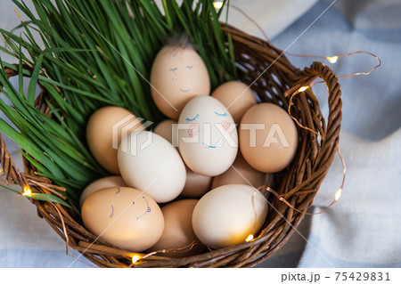 Beautiful painted Easter eggs with a cute face that lie in a wooden basket decorated with warm lights. Easter card, top view. Beautiful painted Easter eggs with a cute face that lie in a wooden basket decorated with warm lights. Easter card, top view. 75429831
