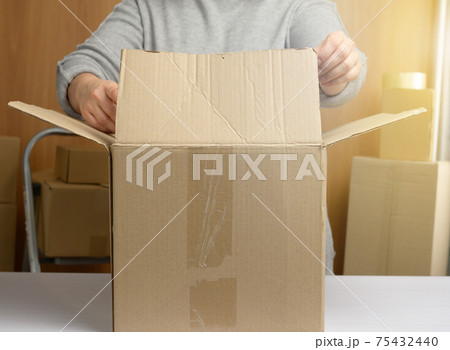 woman in a gray sweater is packing brown cardboard boxes on a white table, behind a stack of boxes woman in a gray sweater is packing brown cardboard boxes on a white table, behind a stack of boxes 75432440