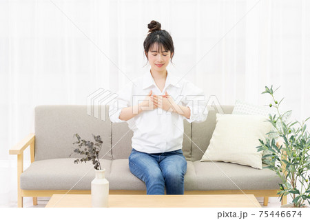 窓際のソファに座って胸に手を置く若い女性 75446674
