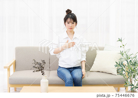 窓際のソファに座って胸に手を置く若い女性 75446676