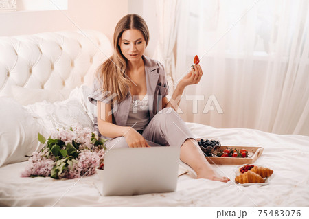 a girl sits in bed in the evening watching a laptop and eating strawberries, a girl in bed eats sweets before going to bed 75483076