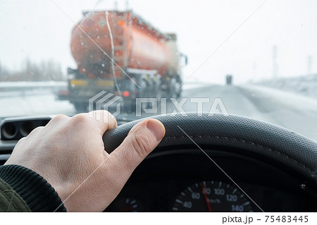 the driver's hand on the steering wheel of the car, which is overtaken on the secondary line by a tanker truck with a large tank of fuel 75483445