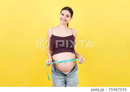 Cropped image of expecting mother in unzipped jeans measuring her pregnant belly with a tape measure at colorful background. Centimeter measurement Cropped image of expecting mother in unzipped jeans measuring her pregnant belly with a tape measure at colorful background. Centimeter measurement 75487545