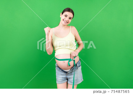 Cropped image of expecting mother in unzipped jeans measuring her pregnant belly with a tape measure at colorful background. Centimeter measurement 75487565