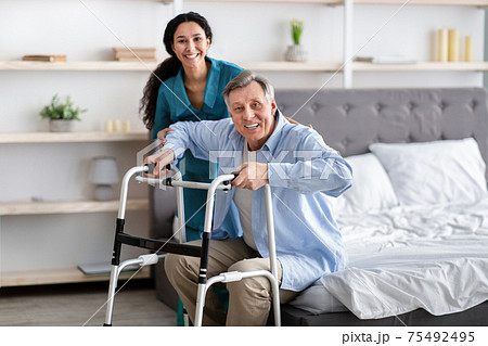 Female nurse helping elderly male with walking frame stand up from bed at home. Professional care for disabled patients Female nurse helping elderly male with walking frame stand up from bed at home. Professional care for disabled patients 75492495