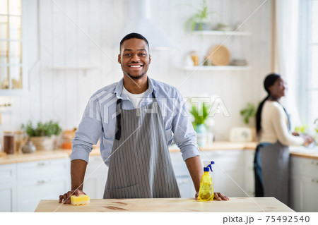 Portrait of black guy in apron and rubber gloves smiling at camera at kitchen, his wife washing dishes on background Portrait of black guy in apron and rubber gloves smiling at camera at kitchen, his wife washing dishes on background 75492540