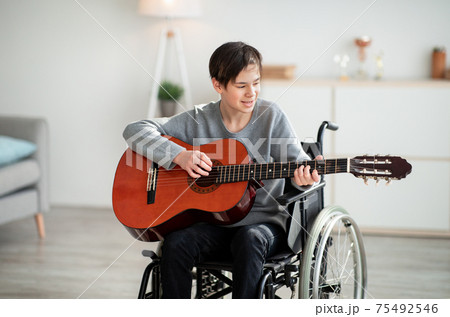 Handicapped teen boy in wheelchair playing guitar and singing indoors. Stay home hobbies 75492546