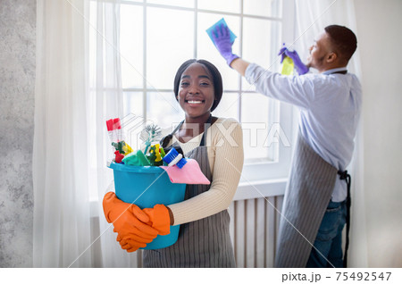 Cheerful black lady holding bucket of cleaning supplies while her husband wiping window at modern flat 75492547