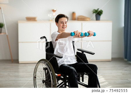 Physical rehabilitation for disabled people. Handicapped teenager in wheelchair making exerises with dumbbells at home Physical rehabilitation for disabled people. Handicapped teenager in wheelchair making exerises with dumbbells at home 75492555