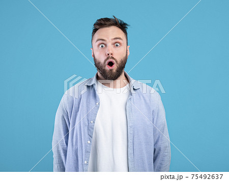 Portrait of stunned young guy opening mouth in shock on blue studio background 75492637