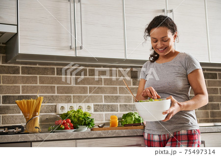 Smiling African American woman stirring vegetable salad with wooden spoon while smiling in kitchen. Houselife and healthy eating concept 75497314