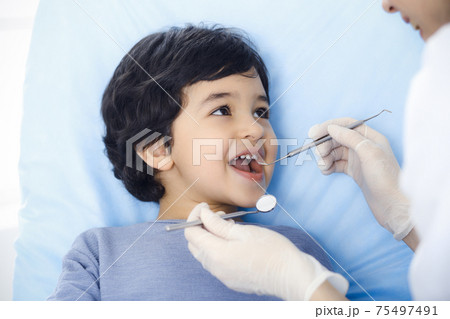Cute arab boy sitting at dental chair with open mouth during oral checking up with doctor. Visiting dentist office. Stomatology concept 75497491