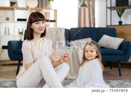 Young Caucasian mother holding hygienic cotton swabs, sitting on the floor in cozy living room with her cute little daughter, wrapped in towel after bathing at home. Hygiene and care concept Young Caucasian mother holding hygienic cotton swabs, sitting on the floor in cozy living room with her cute little daughter, wrapped in towel after bathing at home. Hygiene and care concept 75497859