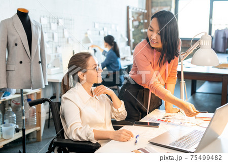 Happy young female designer in wheelchair looking at her Asian colleague pointing at laptop display Happy young female designer in wheelchair looking at her Asian colleague pointing at laptop display 75499482