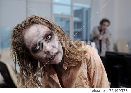 Zombie businesswoman standing in front of camera on background of workplaces and dead male colleague Zombie businesswoman standing in front of camera on background of workplaces and dead male colleague 75499573