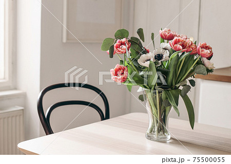 Spring flowers in glass vase on wooden table. Blurred kitchen background with old chair. Bouquet of red tulips, white anemone flowers and eucalyptus branches. Contemporary elegant, Scandinavian 75500055