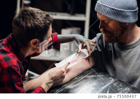 A tattoo artist bandages the young man's hand with a thin layer of petroleum jelly after applying the tattoo. A tattoo artist bandages the young man's hand with a thin layer of petroleum jelly after applying the tattoo. 75500437