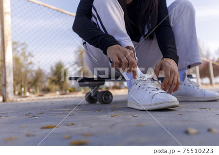 Young women sitting on a surf skate board tying a shoe before play. Young girl long hair sitting and tying a sneaker at public park on morning before play surf skate board 75510223