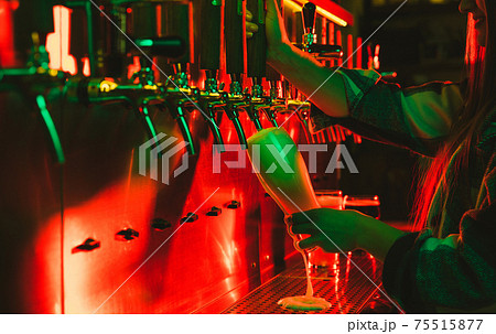Young woman barman pouring a cold frothy beer from tap to glass in neon light. Young woman barman pouring a cold frothy beer from tap to glass in neon light. 75515877